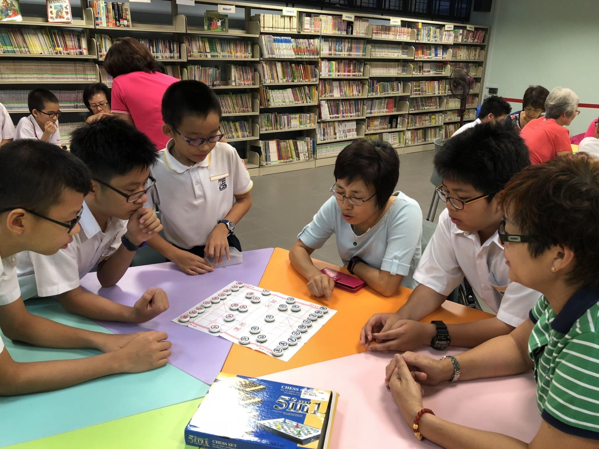 Children and adults gathered around a table playing Chinese chess in a library with full bookshelves in the background.